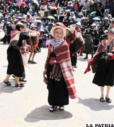 MAMA JILAKATA – MAMA’TAIKA: Es la compañera del jilakata, juntos son autoridades originarias del ayllu, velan por el bienestar de los wawaqallus (comunarios). En el conjunto, éste personaje recae en las promesantes más antiguas que tienen a su cargo a las mujeres que componen los bloques.
Viste con un urku o vestido con alforzas hecha de bayeta de la tierra o tocuyo, en su espalda lleva un aguayo multicolor, para darle varios usos, que lo lleva cruzado desde el hombro hasta la cintura sujeto con un tupu, que es una especie de alfiler cuya cabeza presenta una plancha delgada y redonda trabajada en alto relieve.
A la vez usa una wist’alla o bolsa de aguayo para llevar coca, tejida con lana de oveja que va cruzada a ambos lados del cuerpo.
Asimismo, la inkuña o tarilla es una especie de pañuelo de aguayo anudado en una de sus puntas en la que lleva coca, en su cintura se coloca una faja delgada tejida con lana de oveja.
Tiene un sombrero o tanka fabricado con lana de oveja, calza wikhus o abarcas hechas de goma o cuero forrado con lana negra.